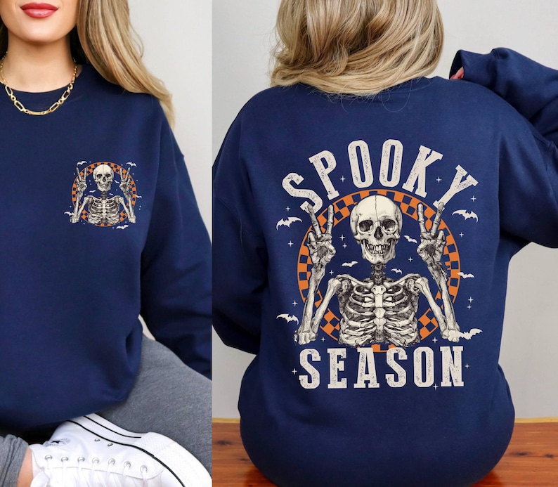 Puede incluir: Sudadera azul marino con un gr&aacute;fico de esqueleto y el texto "Spooky Season" en blanco y naranja. El esqueleto lleva un collar con un signo de paz y tiene los brazos levantados en un gesto de paz.