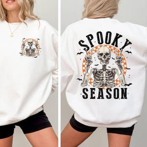 Puede incluir: Sudadera blanca con un gr&aacute;fico de esqueleto en la parte delantera y el texto "Spooky Season" con un gr&aacute;fico de esqueleto en la parte posterior.