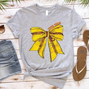 May include: Grey t-shirt with a yellow softball bow and the text "Your Team" printed on the front.