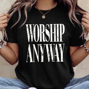 May include: Black t-shirt with the white text "WORSHIP ANYWAY". The shirt has short sleeves and a crew neck. The text has a distressed, vintage style. The shirt is made of a soft material.