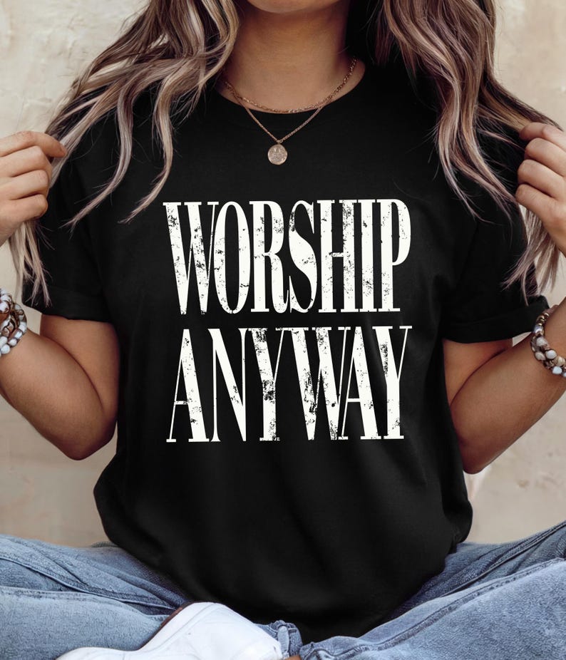 May include: Black t-shirt with the white text "WORSHIP ANYWAY". The shirt has short sleeves and a crew neck. The text has a distressed, vintage style. The shirt is made of a soft material.
