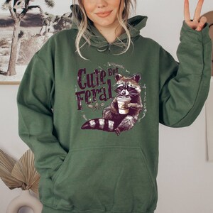 May include: Green hooded sweatshirt with a graphic of a raccoon holding a coffee cup and the text "Cute But Feral".