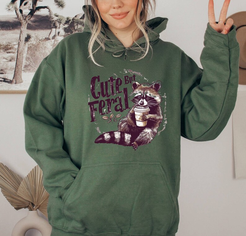 May include: Green hooded sweatshirt with a graphic of a raccoon holding a coffee cup and the text "Cute But Feral".