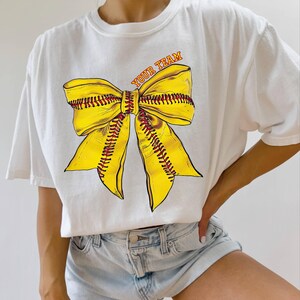 May include: White t-shirt featuring a large yellow softball bow graphic with red stitching and the text "YOUR TEAM" in orange. The bow design is centered on the shirt.