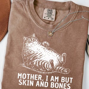 以下が含まれることがあります： リラックスした猫のイラストと「MOTHER, I AM BUT SKIN AND BONES」の文字が入ったエスプレッソカラーのTシャツ。シャツにはComfort Colorsのタグとペンダント付きのシルバーネックレスが付いています。シャツは折りたたまれています。