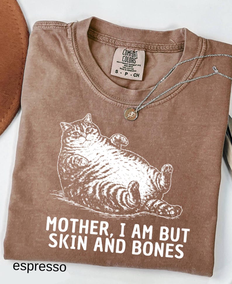 以下が含まれることがあります： リラックスした猫のイラストと「MOTHER, I AM BUT SKIN AND BONES」の文字が入ったエスプレッソカラーのTシャツ。シャツにはComfort Colorsのタグとペンダント付きのシルバーネックレスが付いています。シャツは折りたたまれています。