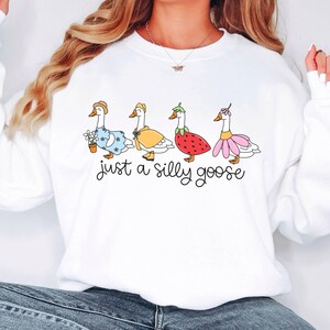 May include: White sweatshirt with the phrase "just a silly goose" in black script. The design features four cartoon geese dressed in various outfits, including a dress, a raincoat, and a strawberry costume.
