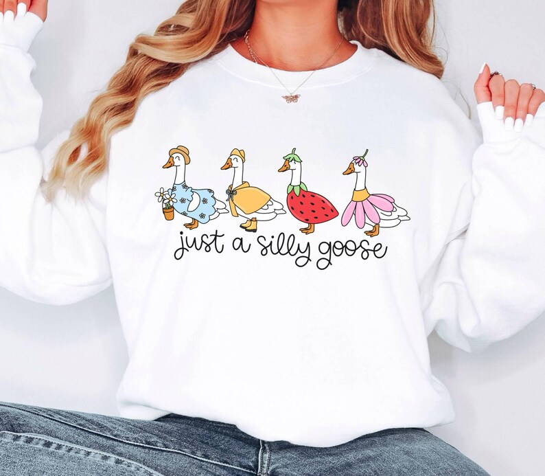 May include: White sweatshirt with the phrase "just a silly goose" in black script. The design features four cartoon geese dressed in various outfits, including a dress, a raincoat, and a strawberry costume.