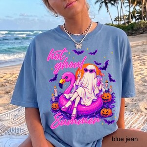 May include: A blue t-shirt featuring a graphic print of a ghost wearing sunglasses and sitting on a pink flamingo pool float. The design includes text that reads hot ghoul Summer, surrounded by purple bats, orange jack-o'-lanterns, and candles.