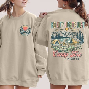 May include: A beige sweatshirt with a graphic of a mountain scene and the text "No City Lights Just Camp Fire Nights" on the back. The front of the sweatshirt has a smaller graphic of a mountain scene.