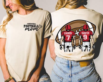 May include: Cream-colored t-shirt with the text "Football Mom" in black script. The back of the shirt features a football graphic with two football players, one with the name "James" and the number 7, and the other with "David" and the number 9.