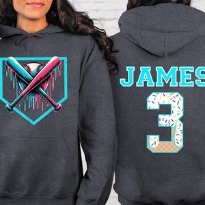 May include: Dark gray hoodie with a baseball-themed graphic on the front, featuring crossed bats and a baseball within a teal outline. The back displays the name "JAMES" and the number "3" with a sprinkle-covered design.