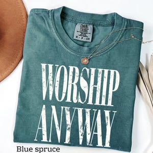 May include: A blue spruce Comfort Colors t-shirt with the words "WORSHIP ANYWAY" in white block letters. A gold necklace with a pendant rests on the shirt. The shirt is folded neatly.