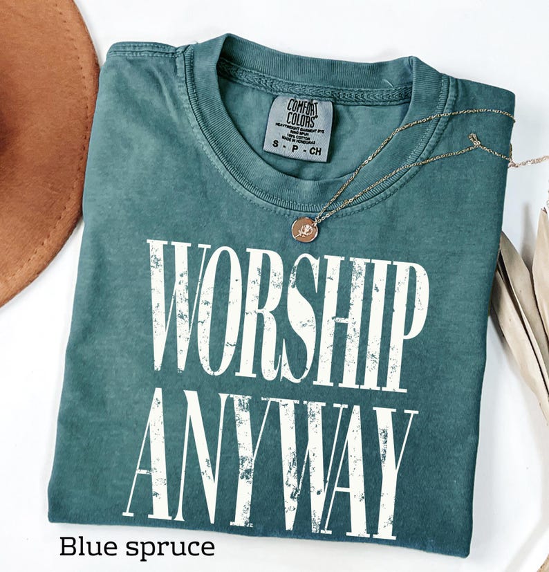 May include: A blue spruce Comfort Colors t-shirt with the words "WORSHIP ANYWAY" in white block letters. A gold necklace with a pendant rests on the shirt. The shirt is folded neatly.