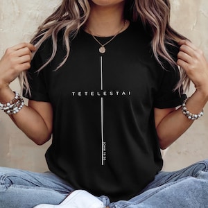 May include: Black t-shirt with a white cross design. The word "TETELESTAI" is printed across the top of the cross, with "JOHN 19:30" printed vertically below. The shirt is a classic crew neck style.