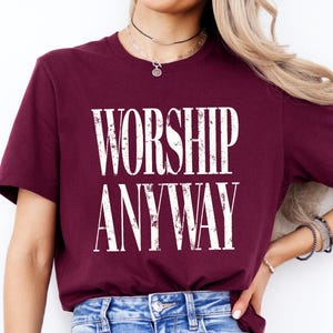 May include: Burgundy crew neck t-shirt with the words "WORSHIP ANYWAY" printed in a distressed white font. The shirt is made of a soft material and is suitable for casual wear.