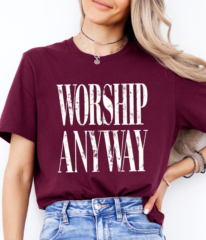 May include: Burgundy crew neck t-shirt with the words "WORSHIP ANYWAY" printed in a distressed white font. The shirt is made of a soft material and is suitable for casual wear.