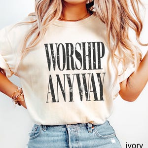 May include: Ivory-colored t-shirt with the words "WORSHIP ANYWAY" printed in bold, black letters. The shirt is a light cream color and has short sleeves. The text is centered on the front of the shirt.