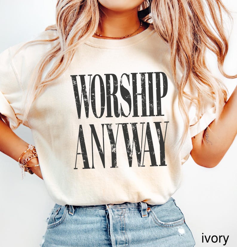 May include: Ivory-colored t-shirt with the words "WORSHIP ANYWAY" printed in bold, black letters. The shirt is a light cream color and has short sleeves. The text is centered on the front of the shirt.
