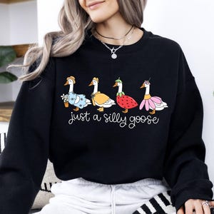 May include: Black sweatshirt featuring a whimsical illustration of four geese, each adorned with unique outfits and accessories. The phrase "just a silly goose" is written in cursive below the geese. A fun and playful design.