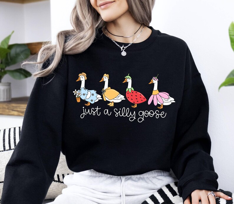 May include: Black sweatshirt featuring a whimsical illustration of four geese, each adorned with unique outfits and accessories. The phrase "just a silly goose" is written in cursive below the geese. A fun and playful design.