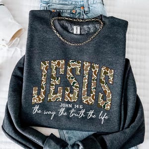May include: Dark gray sweatshirt with the word "JESUS" in a camouflage pattern. The text "JOHN 14:6" and "the way the truth the life" are printed below the word "JESUS". A gold chain is draped around the collar.