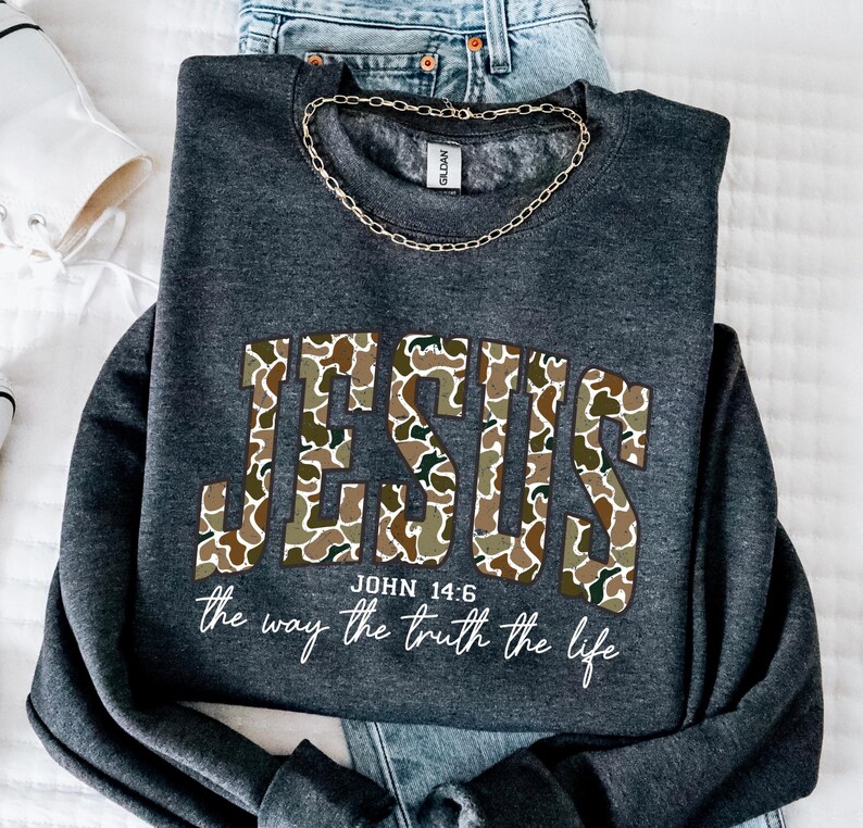 May include: Dark gray sweatshirt with the word "JESUS" in a camouflage pattern. The text "JOHN 14:6" and "the way the truth the life" are printed below the word "JESUS". A gold chain is draped around the collar.