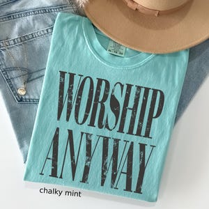 May include: A chalky mint-colored t-shirt with the words "WORSHIP ANYWAY" printed in distressed black lettering. The shirt is folded and placed on top of blue jeans and a tan hat.