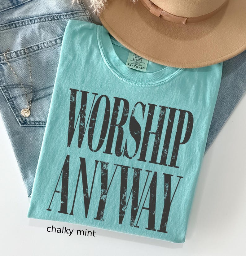 May include: A chalky mint-colored t-shirt with the words "WORSHIP ANYWAY" printed in distressed black lettering. The shirt is folded and placed on top of blue jeans and a tan hat.