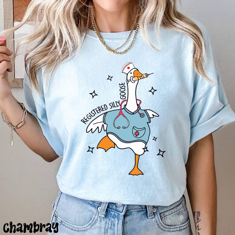 K&ouml;nnte beinhalten: Hellblaues T-Shirt mit einer Zeichnung einer Gans, die eine Krankenschwesterm&uuml;tze und einen Kittel tr&auml;gt. Die Gans h&auml;lt eine Spritze und der Text "Registered Silly Goose" ist auf dem T-Shirt aufgedruckt.