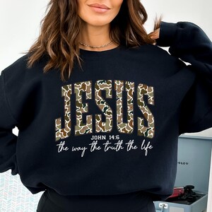 May include: Black sweatshirt with the word "JESUS" in camouflage print across the chest. Below the word "JESUS" is the text "JOHN 14:6" and the phrase "the way the truth the life" in a white script font.