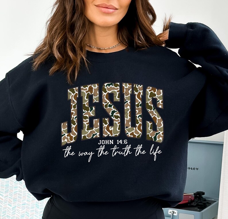 May include: Black sweatshirt with the word "JESUS" in camouflage print across the chest. Below the word "JESUS" is the text "JOHN 14:6" and the phrase "the way the truth the life" in a white script font.