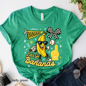 May include: Kelly green t-shirt with a graphic of a smiling banana wearing sunglasses, holding a baseball bat, and a baseball. The text reads "Bananas" and "Let's Go Bananas". A checkered bow and a foam finger are also featured.