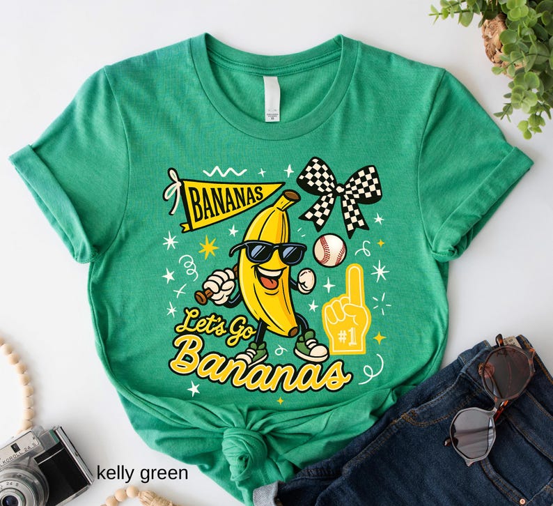May include: Kelly green t-shirt with a graphic of a smiling banana wearing sunglasses, holding a baseball bat, and a baseball. The text reads "Bananas" and "Let's Go Bananas". A checkered bow and a foam finger are also featured.