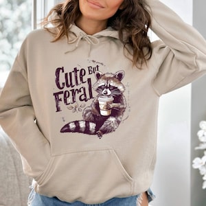 May include: A beige hoodie with a graphic of a raccoon holding a coffee cup. The text "Cute But Feral" is printed on the hoodie.