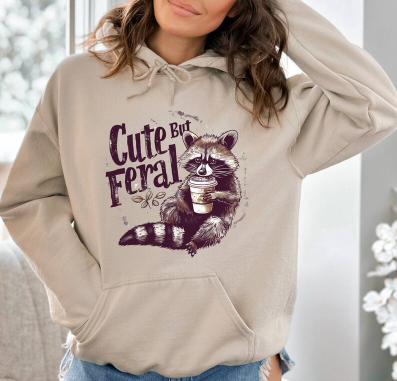 May include: A beige hoodie with a graphic of a raccoon holding a coffee cup. The text "Cute But Feral" is printed on the hoodie.