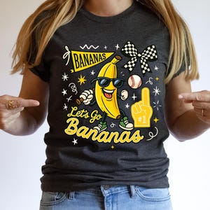 May include: Dark gray t-shirt with a cartoon banana wearing sunglasses and sneakers. The shirt features the text "BANANAS" on a banner and "Let's Go Bananas" in yellow script, along with baseball and foam finger graphics.