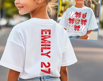 Può includere: Maglietta bianca con testo rosso sul retro che dice "Emily 27". La parte anteriore della maglietta ha un design di palle da baseball, mazze, fiocchi e il testo "Your Team Name".