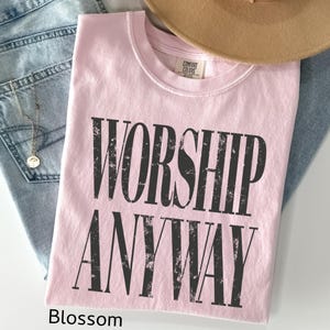 May include: A light pink t-shirt with the words "WORSHIP ANYWAY" printed in distressed black lettering. The shirt is folded and rests on a pair of blue jeans, with a tan hat and a gold necklace visible.