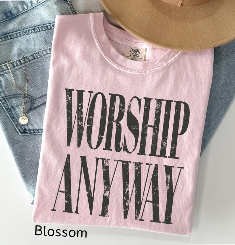 May include: A light pink t-shirt with the words "WORSHIP ANYWAY" printed in distressed black lettering. The shirt is folded and rests on a pair of blue jeans, with a tan hat and a gold necklace visible.