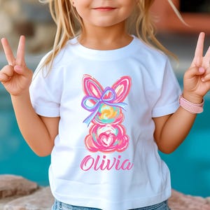 May include: White t-shirt with a colorful abstract bunny design and the name "Olivia" printed in pink.