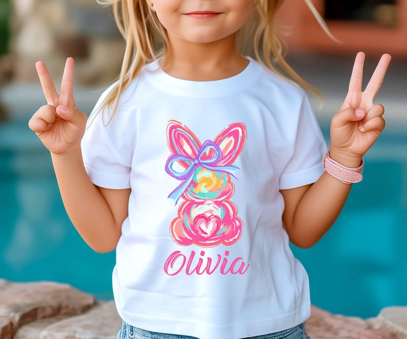 May include: White t-shirt with a colorful abstract bunny design and the name "Olivia" printed in pink.