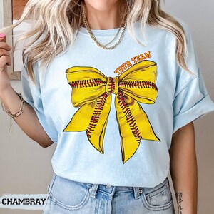 May include: Light blue t-shirt featuring a large yellow softball bow graphic with red stitching and the text "YOUR TEAM" in orange. The shirt is made of soft material and is suitable for casual wear. The word "CHAMBRAY" is printed at the bottom left.