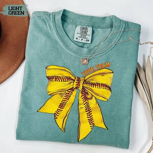 May include: Light green t-shirt with a large yellow softball bow graphic and the text "YOUR TEAM" in orange. The shirt has a crew neck and short sleeves. A silver necklace with a pendant is draped on the shirt.