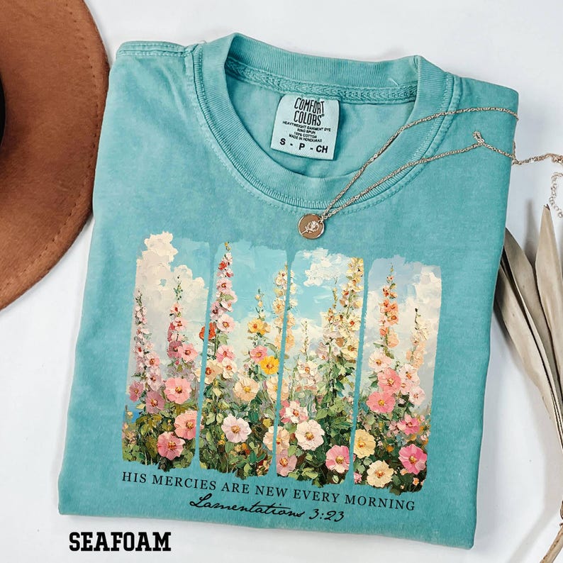 May include: Seafoam green Comfort Colors t-shirt with a floral print. The graphic features colorful flowers against a blue sky, with the text "HIS MERCIES ARE NEW EVERY MORNING Lamentations 3:23". A gold necklace rests on the shirt.