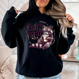 May include: Black hooded sweatshirt with a graphic of a raccoon holding a coffee cup and the text "Cute But Feral".