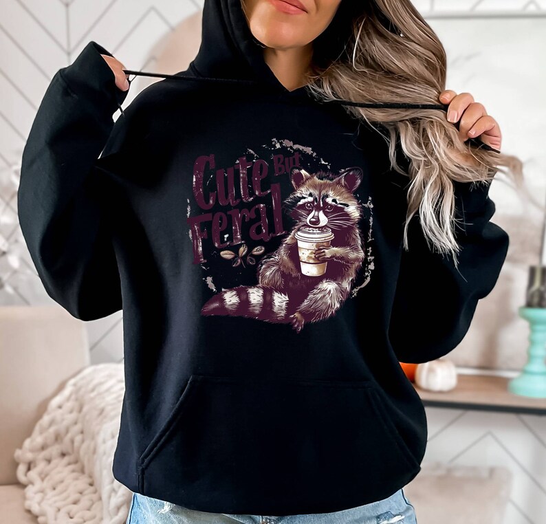 May include: Black hooded sweatshirt with a graphic of a raccoon holding a coffee cup and the text "Cute But Feral".