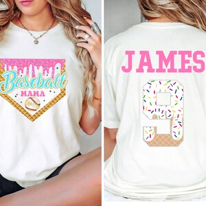 May include: White t-shirt with a pink, blue, and yellow baseball-themed graphic that says "Baseball Mama" on the front. The back of the shirt has the name "JAMES" in pink letters and a number "9" with sprinkles on it.