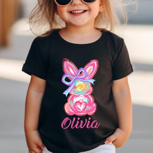 May include: Black t-shirt with a colorful Easter bunny graphic and the name "Olivia" printed on the front.
