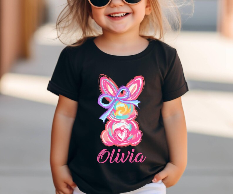 May include: Black t-shirt with a colorful Easter bunny graphic and the name "Olivia" printed on the front.